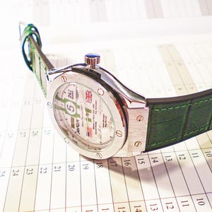 May include: A silver-toned wristwatch with a green leather band. The watch face displays the number 5, the Union Jack, and other text. The watch is resting on a calendar.
