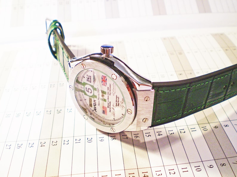 May include: A silver-toned wristwatch with a green leather band. The watch face displays the number 5, the Union Jack, and other text. The watch is resting on a calendar.
