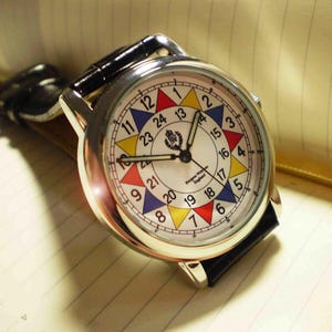 May include: A silver wristwatch with a white face and black leather band. The face has a colorful 24-hour clock design with red, yellow, blue, and green accents. The watch face also includes the text "Burgess Watch Co. England".
