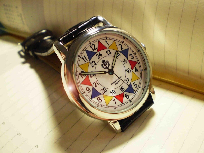 May include: A silver wristwatch with a white face and black leather band. The face has a colorful 24-hour clock design with red, yellow, blue, and green accents. The watch face also includes the text "Burgess Watch Co. England".