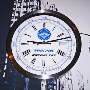 Könnte beinhalten: Ein weißes Zifferblatt mit schwarzen römischen Ziffern für die Stunden und schwarzen Zeigern. Das Zifferblatt hat ein blaues und weißes Pan Am-Logo und den Text "PAN AM BOEING 747". Die Uhr befindet sich in einem silbernen Metallrahmen.