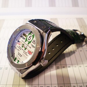 May include: A silver-toned wristwatch with a white face featuring green and black accents, the number 5, and the Union Jack. The watch has a dark green leather strap and is set against a calendar.