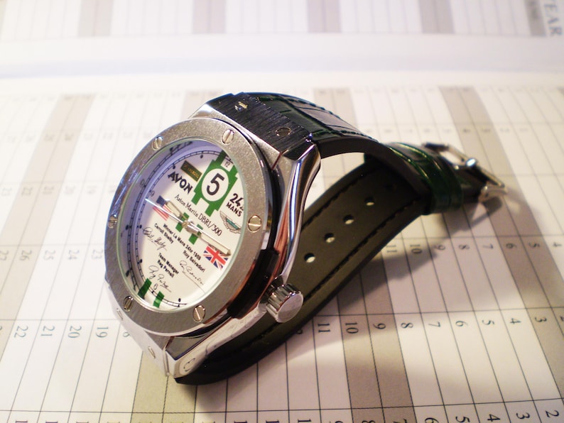 May include: A silver-toned wristwatch with a white face featuring green and black accents, the number 5, and the Union Jack. The watch has a dark green leather strap and is set against a calendar.