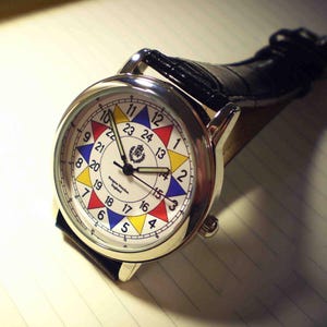 May include: A silver wristwatch with a black leather band. The watch face has a white background with a colorful circular design and black hands. The numbers on the watch face are in black and white.