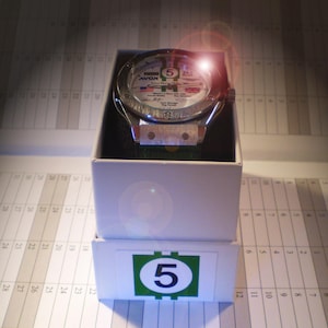 May include: A silver-toned wristwatch with a white face, featuring the number 5 and various text details, presented in a white box. The watch has a green strap and is displayed on a calendar-style background. The box has a green and white label with the number 5.