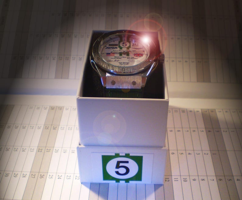 May include: A silver-toned wristwatch with a white face, featuring the number 5 and various text details, presented in a white box. The watch has a green strap and is displayed on a calendar-style background. The box has a green and white label with the number 5.