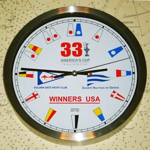 May include: A round wall clock with a silver-colored metal frame. The clock face is white with black hands and features the text "33 AMERICA'S CUP VALENCIA" in red. It also displays nautical flags and the text "WINNERS USA".