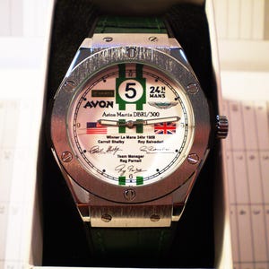 May include: A silver wristwatch with a white face featuring green and black accents, the number 5, and the text "24 H MANS". The watch includes the text "AVON Aston Martin DBR1/300" and the flags of the United States and the United Kingdom.