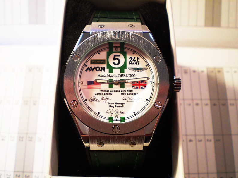 May include: A silver wristwatch with a white face featuring green and black accents, the number 5, and the text "24 H MANS". The watch includes the text "AVON Aston Martin DBR1/300" and the flags of the United States and the United Kingdom.
