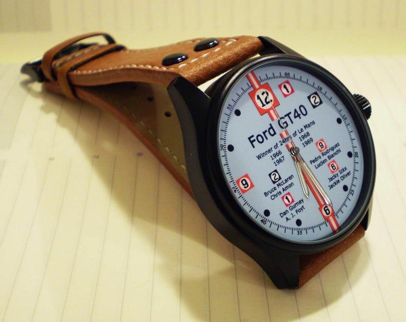May include: A black wristwatch with a brown leather strap. The watch face is white with black numbers and a red stripe. The text "Ford GT40" is printed on the watch face, along with the words "Winner of 24hrs of Le Mans" and the years 1966, 1967, 1968, and 1969. The names of the drivers who won the race in those years are also listed on the watch face.