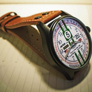 K&ouml;nnte beinhalten: Eine schwarz-braune Armbanduhr mit wei&szlig;em Zifferblatt und gr&uuml;nen und roten Akzenten. Das Zifferblatt der Uhr zeigt den Text "Avon 24H Mans" und "Winner Le Mans 1923 Carroll Shelby".