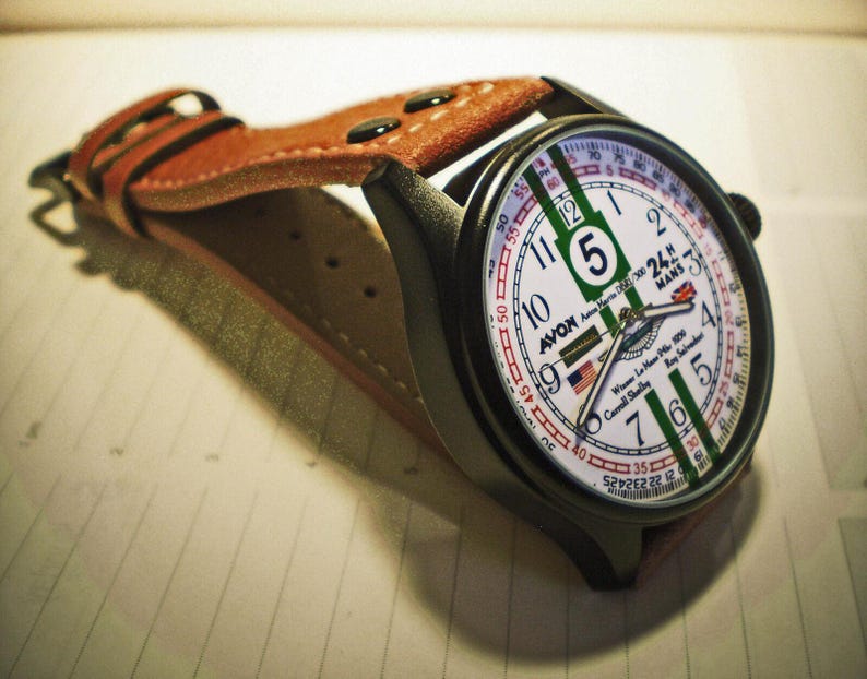 K&ouml;nnte beinhalten: Eine schwarz-braune Armbanduhr mit wei&szlig;em Zifferblatt und gr&uuml;nen und roten Akzenten. Das Zifferblatt der Uhr zeigt den Text "Avon 24H Mans" und "Winner Le Mans 1923 Carroll Shelby".