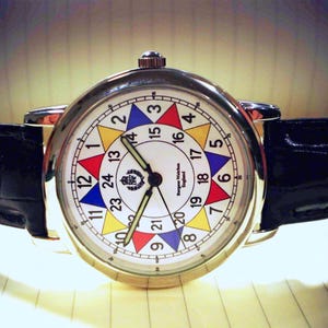 May include: A white analog wristwatch with a black leather band. The watch face has a colorful design with numbers 1 through 24 arranged in a circle. The watch face also includes the text "Ingersoll Watches England".