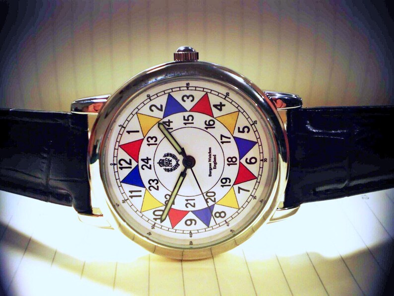 May include: A white analog wristwatch with a black leather band. The watch face has a colorful design with numbers 1 through 24 arranged in a circle. The watch face also includes the text "Ingersoll Watches England".