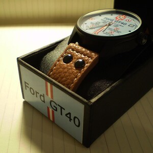 May include: A black watch with a brown leather strap and a white face. The watch face has the text "Ford GT40" on it. The watch is in a black box with the text "Ford GT40" on it.