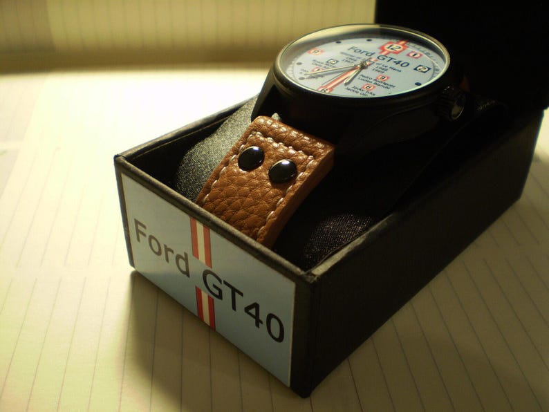 May include: A black watch with a brown leather strap and a white face. The watch face has the text "Ford GT40" on it. The watch is in a black box with the text "Ford GT40" on it.