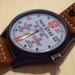 May include: A black and brown wristwatch with a white face and black hands. The face of the watch has the text "Ford GT40" and "Winner of 24hrs of Le Mans" with the years 1966, 1967, and 1969. The watch face also has the numbers 1, 2, 6, 9, and 12.