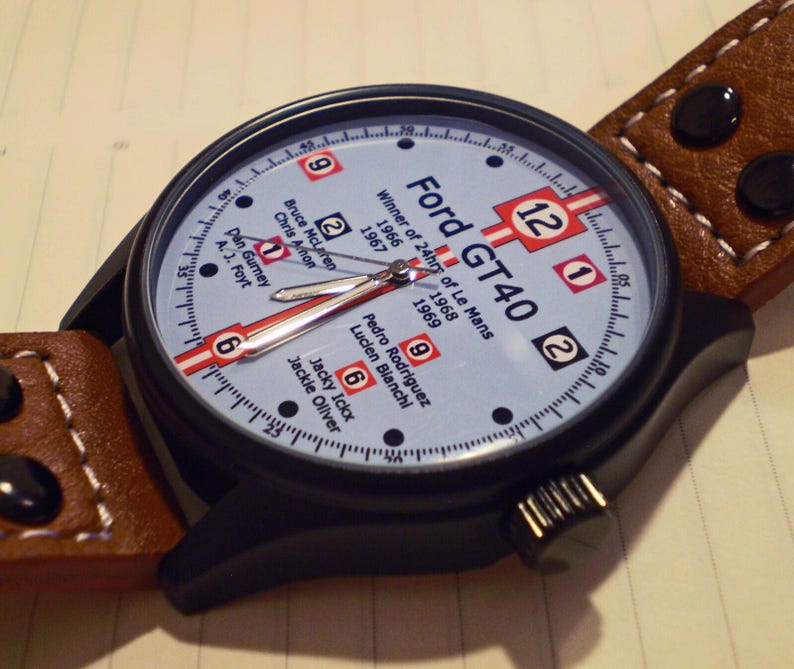 May include: A black and brown wristwatch with a white face and black hands. The face of the watch has the text "Ford GT40" and "Winner of 24hrs of Le Mans" with the years 1966, 1967, and 1969. The watch face also has the numbers 1, 2, 6, 9, and 12.