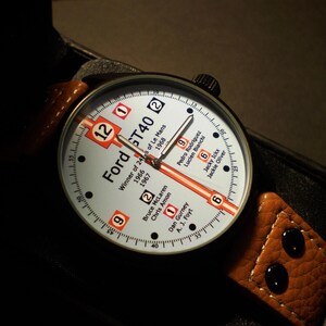 May include: A white and orange wristwatch with a black leather strap. The watch face features the text "Ford GT40" and lists the winners of the 24 Hours of Le Mans from 1966 to 1968.