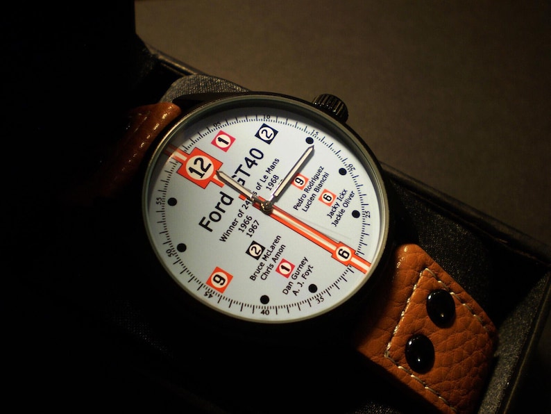 May include: A white and orange wristwatch with a black leather strap. The watch face features the text "Ford GT40" and lists the winners of the 24 Hours of Le Mans from 1966 to 1968.