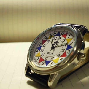 May include: A silver wristwatch with a white face and a black leather band. The face has a colorful design with numbers 1 through 24 around the edge. The watch has a small logo in the center of the face.
