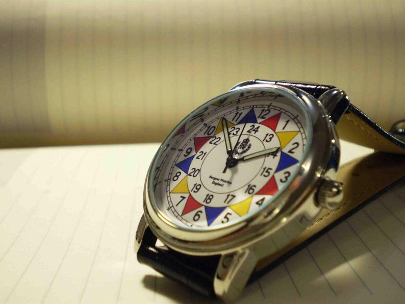 May include: A silver wristwatch with a white face and a black leather band. The face has a colorful design with numbers 1 through 24 around the edge. The watch has a small logo in the center of the face.