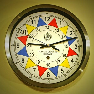 Peut inclure: Un cadran d'horloge rond, blanc, avec des aiguilles noires et une bordure noire. Le cadran est divisé en 12 sections, chacune d'une couleur différente : rouge, jaune, bleu et blanc. Le cadran comporte également les chiffres de 1 à 12, et les mots "BURGESS CLOCK CO ENGLAND" sont imprimés en noir. L'horloge est entourée d'un cadre en métal argenté.