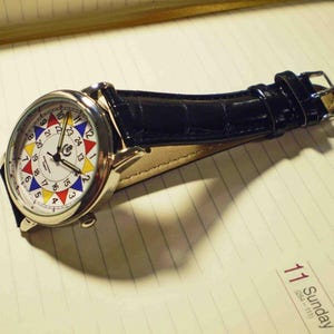 May include: A silver wristwatch with a white face and a black leather band. The face has a colorful circular design with numbers 1 through 24 around the edge. The watch is lying on a lined notebook.