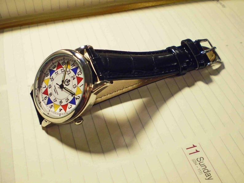May include: A silver wristwatch with a white face and a black leather band. The face has a colorful circular design with numbers 1 through 24 around the edge. The watch is lying on a lined notebook.