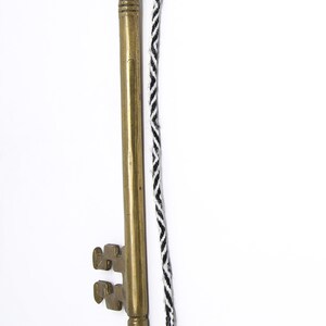May include: A large, antique-looking brass key with a decorative keyhole and a long, black and white braided cord attached to the top.
