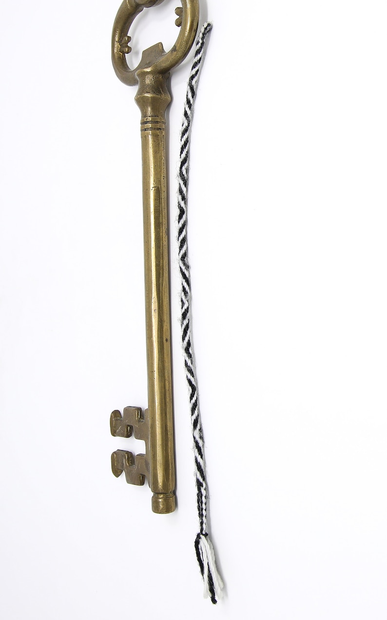 May include: A large, antique-looking brass key with a decorative keyhole and a long, black and white braided cord attached to the top.