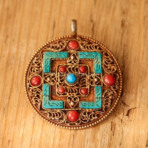 Może przedstawiać: Okrągły wisiorek z filigranowym wzorem w kolorze złotym i czerwonymi i turkusowymi kamieniami. Wisiorek ma kwadratowy kształt w środku z turkusowym kamieniem pośrodku.