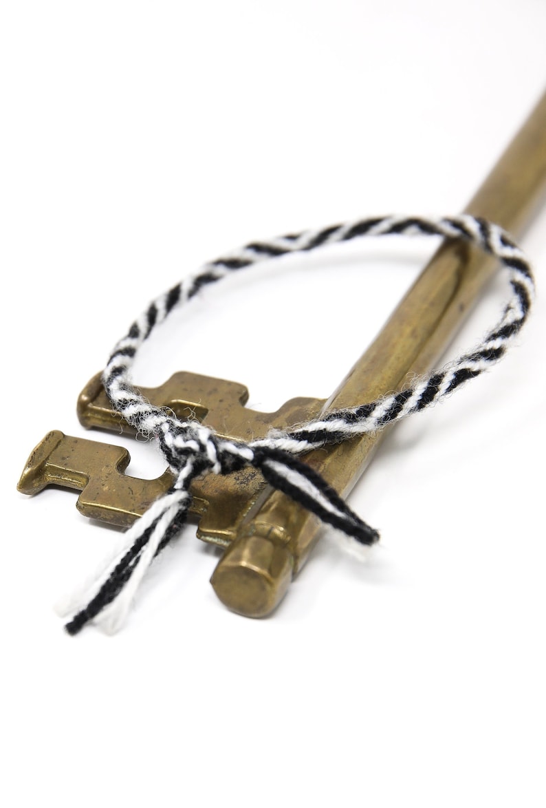 May include: A brass key with a black and white braided cord tied around it. The cord is tied in a knot at the top of the key and has two loose ends.