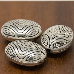 May include: Three silver oval-shaped beads with a geometric pattern. The beads have a textured surface and are arranged on a wooden surface.