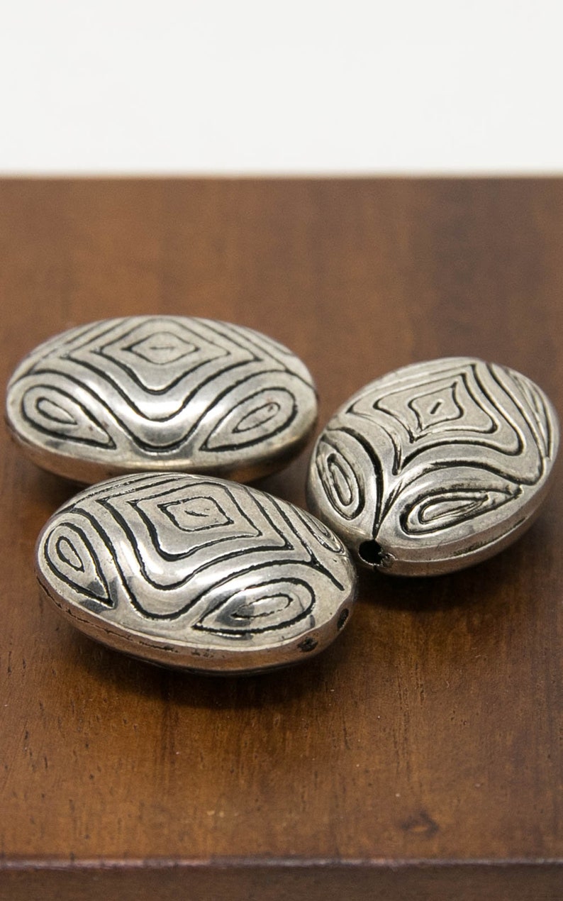 May include: Three silver oval-shaped beads with a geometric pattern. The beads have a textured surface and are arranged on a wooden surface.