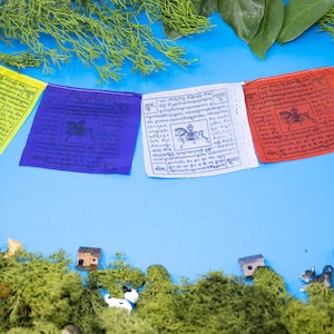 May include: A set of colorful prayer flags with Tibetan writing and images of a horse. The flags are hanging against a blue background with green foliage and miniature houses and animals.