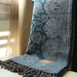 May include: A teal blue paisley patterned scarf with a gray fringe. The scarf is draped over a chair and has a tag that reads "The Little Tibet London".