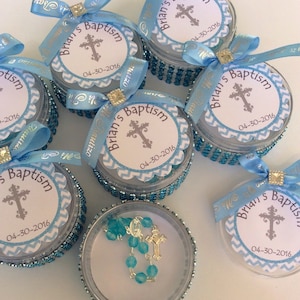 May include: Small, clear containers with blue beaded trim and lids. Each lid has a white label with a cross and the text "Brian's Baptism 04-30-2016". Blue ribbons and bows adorn the containers, some with additional text.