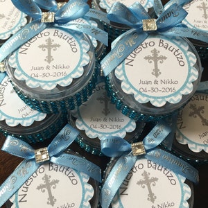 May include: Several small, clear containers with blue ribbon bows and sparkling embellishments. Each container has a circular label with a cross, the words "Nuestro Bautizo", and the names "Juan & Nikko" with the date 04-30-2016.
