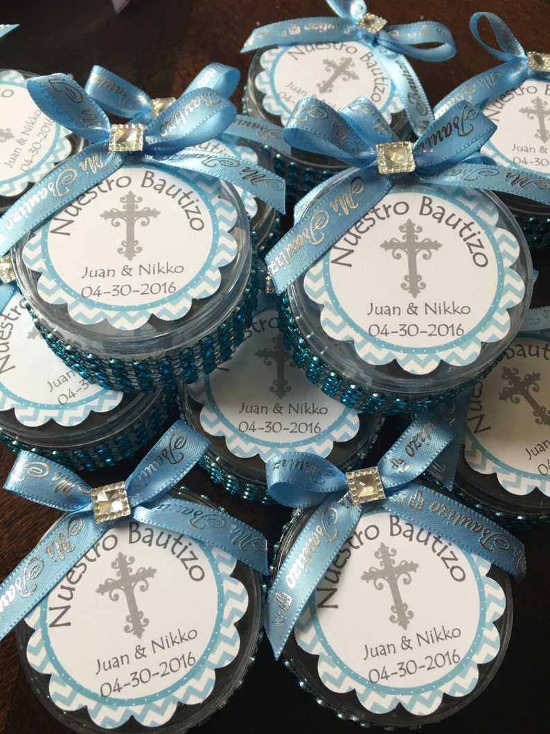 May include: Several small, clear containers with blue ribbon bows and sparkling embellishments. Each container has a circular label with a cross, the words "Nuestro Bautizo", and the names "Juan & Nikko" with the date 04-30-2016.