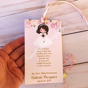 May include: A pink and white bookmark with a floral design and a cartoon girl in a white dress. The bookmark has a gold ribbon and the text "God. Hy ge ardian dear To whom God's love Commits me here. Ever this day be at my side to light and guard. to rule and guide. My First Holy Communion Valerie Vazquez April 28, 2018".
