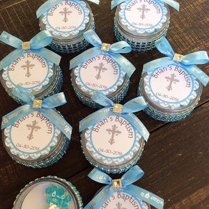 May include: A collection of small, round containers with blue rhinestone trim, each topped with a light blue ribbon bow. The lids feature a circular label with a cross and the text "Brian's Baptism" and the date 04-30-2016.