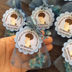 May include: Blue and white favor bags with a blue and white beaded rosary inside. Each bag has a round tag with a picture of a praying angel and the text "Thank you for making my baptism special Eli Maximus 07/16/2016".