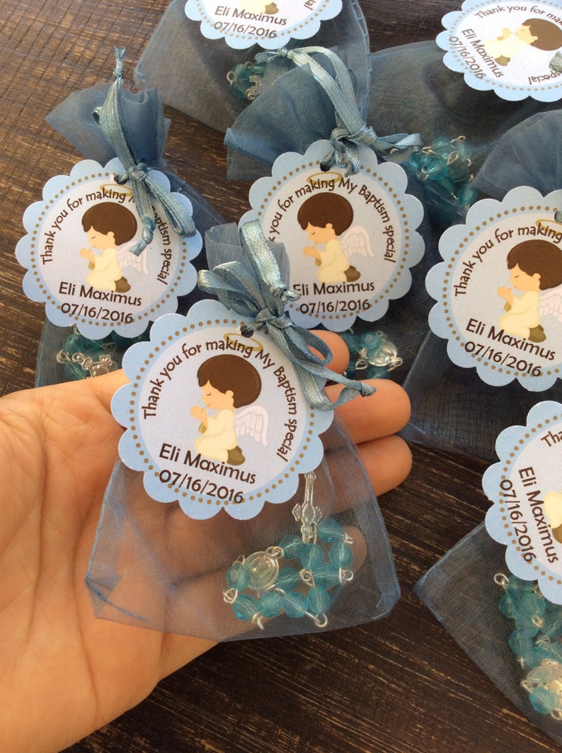 May include: Blue and white favor bags with a blue and white beaded rosary inside. Each bag has a round tag with a picture of a praying angel and the text "Thank you for making my baptism special Eli Maximus 07/16/2016".