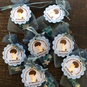 May include: Blue organza bags filled with blue and silver rosary beads and tied with a blue ribbon. Each bag has a round, scalloped, light blue tag with a brown illustration of a boy angel with the text "Thank you for making my baptism special" and "Eli Maximus 07/16/2016".