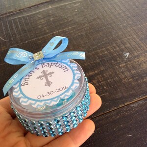 May include: A clear, round container decorated with blue rhinestones and a light blue ribbon. The container has a label that reads "Brian's Baptism" with a cross and the date 04-30-2016. The container is a party favor.