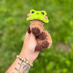 K&ouml;nnte beinhalten: Ein handgefertigtes H&auml;keltier in Form eines braunen B&auml;ren mit einem limettengr&uuml;nen Froschhut. Der B&auml;r hat dunkelbraune Augen und eine hellbraune Schnauze. Das Kuscheltier wird vor einem verschwommenen gr&uuml;nen Hintergrund gehalten.