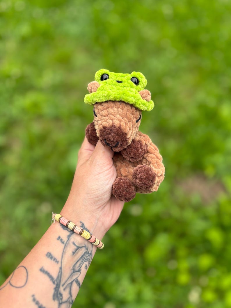 K&ouml;nnte beinhalten: Ein handgefertigtes H&auml;keltier in Form eines braunen B&auml;ren mit einem limettengr&uuml;nen Froschhut. Der B&auml;r hat dunkelbraune Augen und eine hellbraune Schnauze. Das Kuscheltier wird vor einem verschwommenen gr&uuml;nen Hintergrund gehalten.