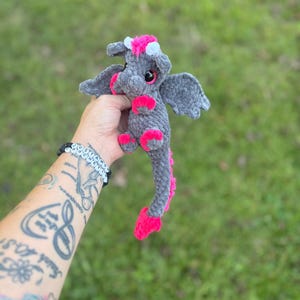 May include: A handmade gray crochet dragon with pink accents. The dragon has small wings, pink feet, and a pink spine. It has a pink and white flower crown. The dragon is held in a person's hand against a green background.