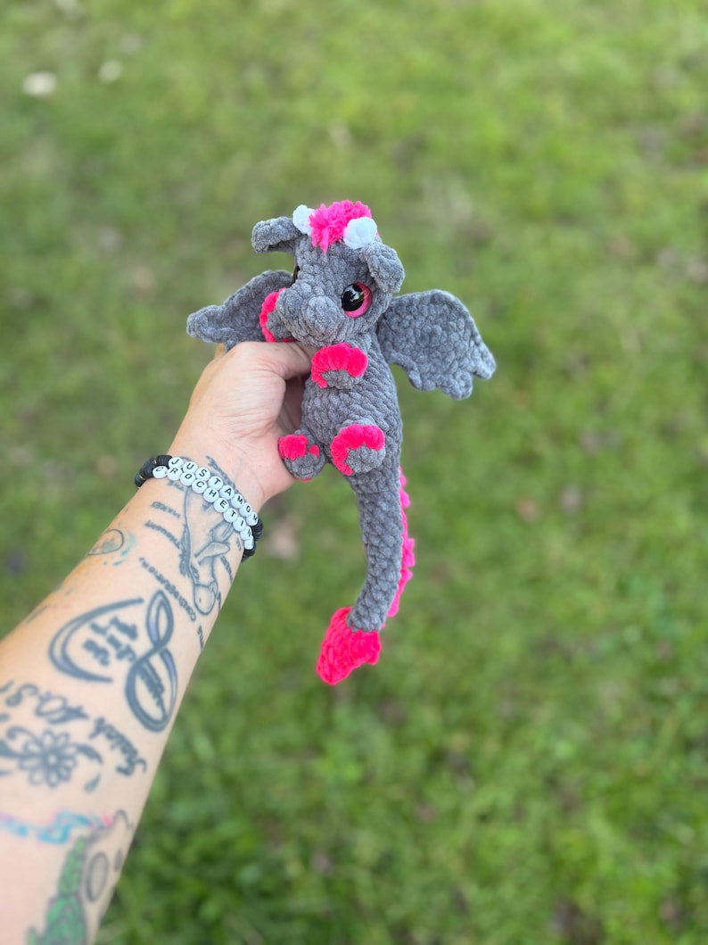 May include: A handmade gray crochet dragon with pink accents. The dragon has small wings, pink feet, and a pink spine. It has a pink and white flower crown. The dragon is held in a person's hand against a green background.